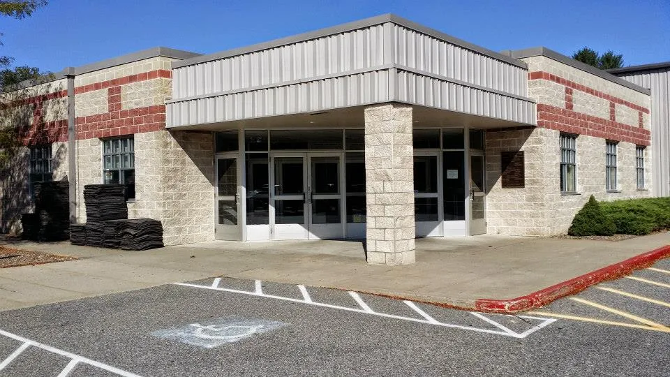 Front entrance of the tan and red brick building, with a canopied entryway, glass double doors and an accessible parking space in the foreground. — RoseGarden Ice Arena