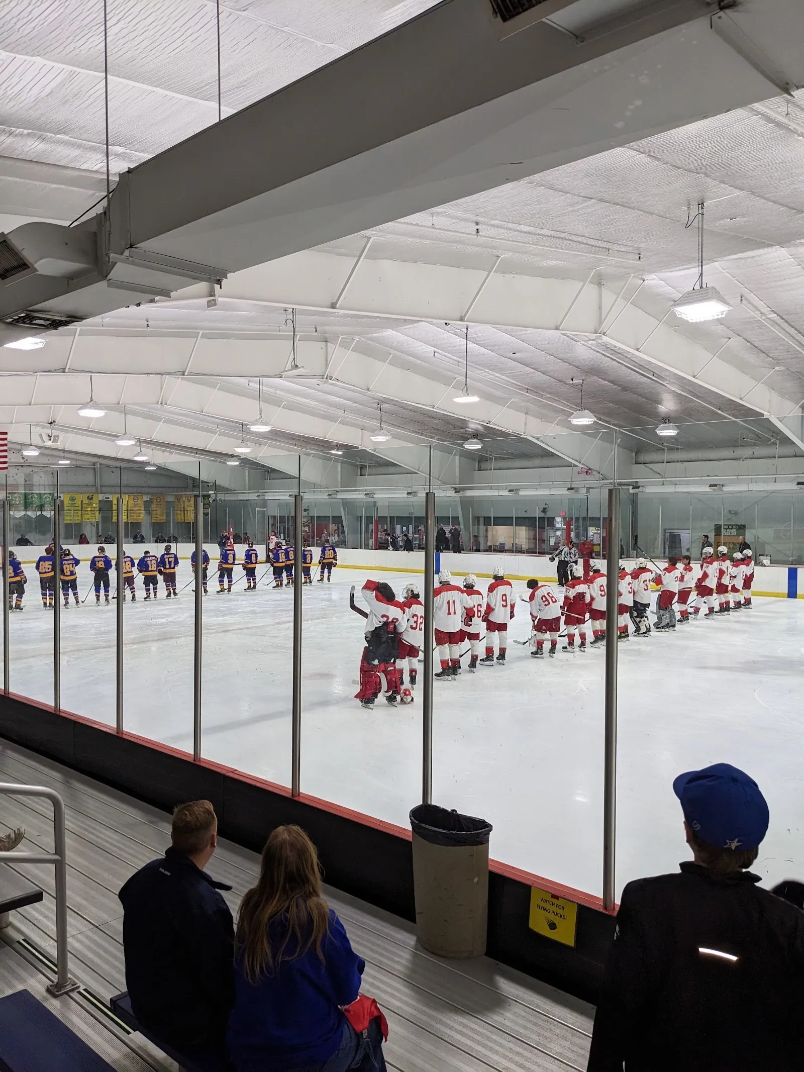 A row of players lined up along the blue line far across the sheet, seen over the glass under an arched white roof with a flag on the far wall. — RoseGarden Ice Arena