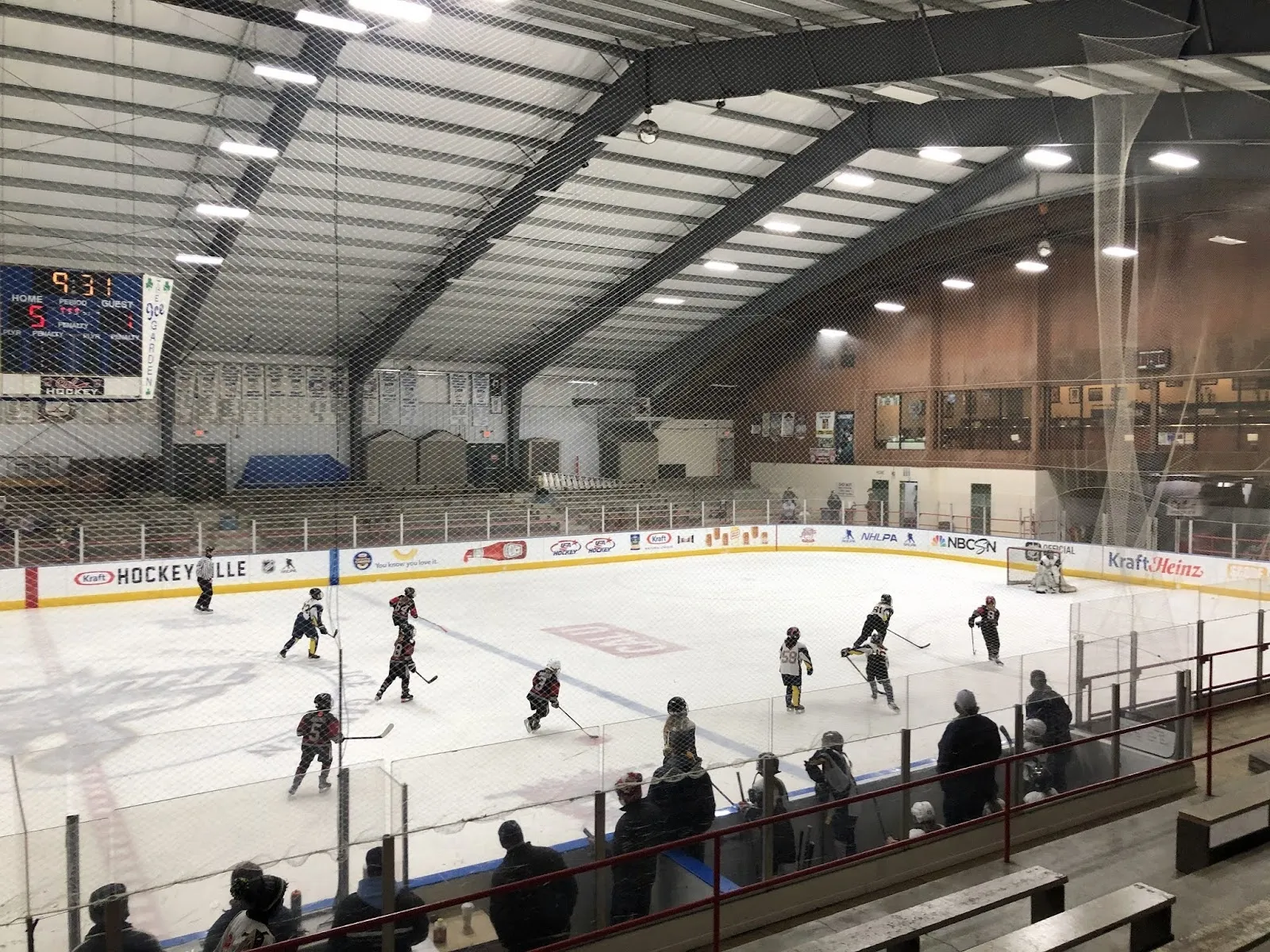 A game seen from the stands, players spread across the ice with a scoreboard at the left and a long dark roof over the rink boards. — Rostraver Ice Garden