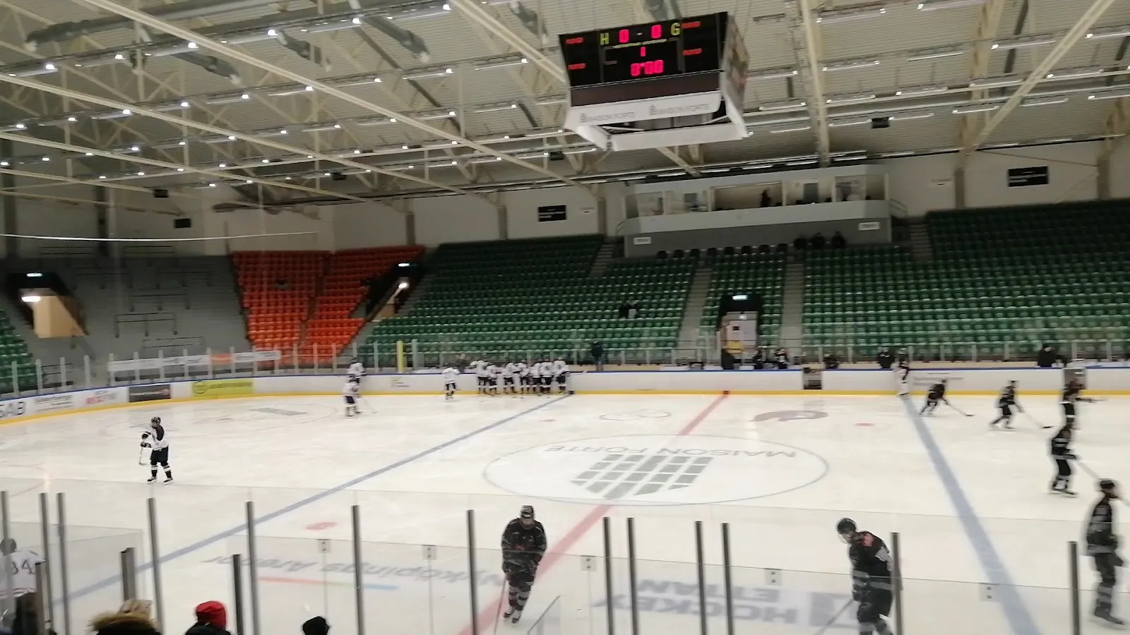 En match på isen i Rosvalla i Nyköping, spelare över rinken under läktarna med gröna säten och resultattavlan. – Rosvalla ishallar