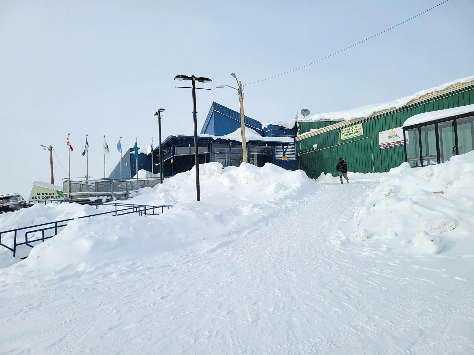 The entrance to the Midnight Sun Complex in Inuvik in winter, with the flags, snowbanks and the green arena wall carrying a sign for the Roy Sugloo Ipana Memorial Arena. — Roy "Sugloo" Ipana Arena