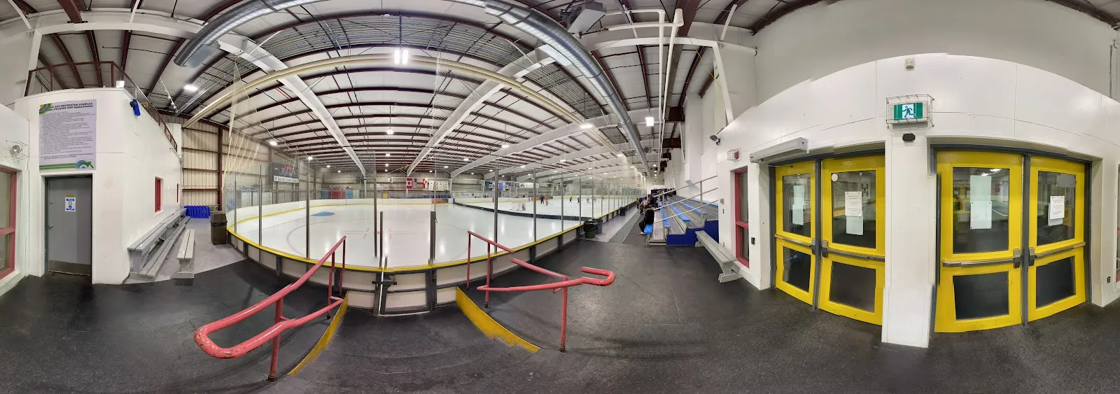A wide panorama inside the Inuvik arena along the boards, with the ice surface, the pink and yellow gate rails and the yellow entrance doors. — Roy "Sugloo" Ipana Arena