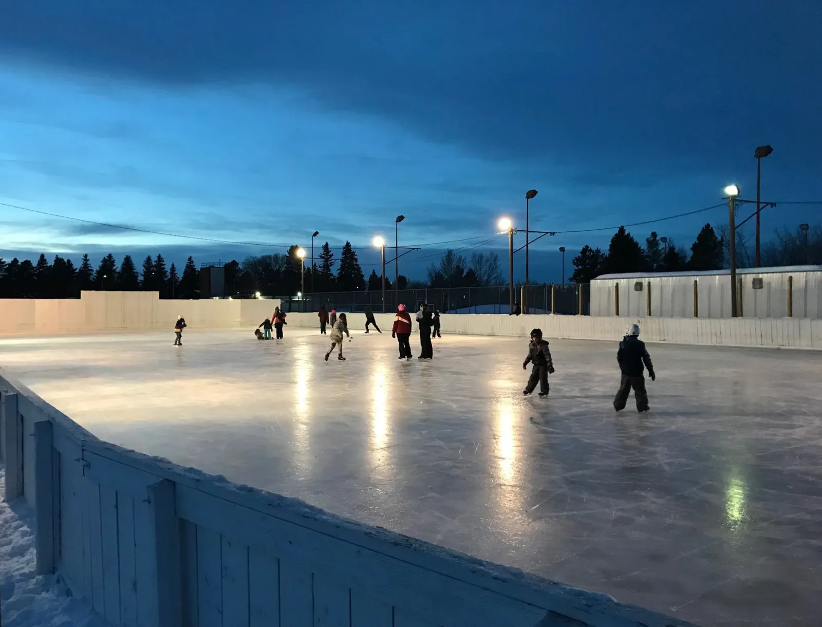 The boarded Royal Gardens community rink at dusk, with skaters at a distance under the floodlights — Royal Gardens Community League Rink