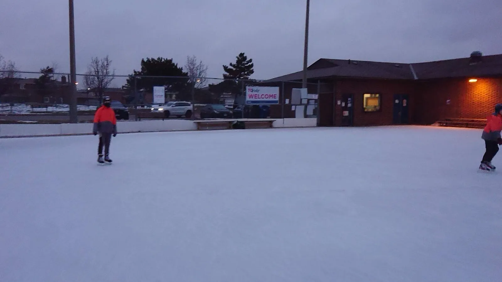 The Royalcrest Park outdoor rink at dusk with the park building behind and skaters at the edge of the ice — Royalcrest Park Outdoor Ice Rink