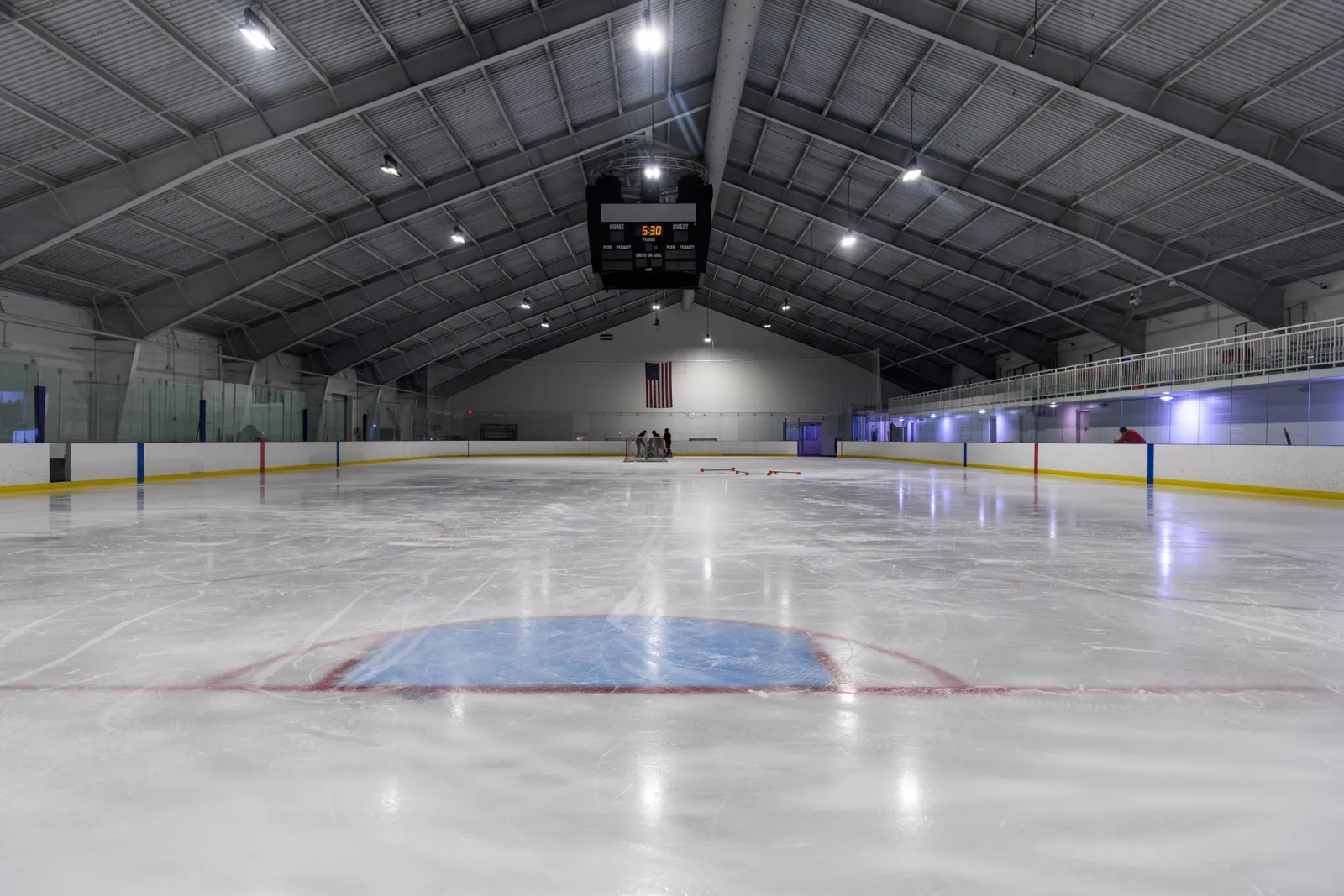 An empty NHL-sized ice sheet shot from ice level toward the far end, under a vaulted white roof, with a hanging scoreboard, an American flag, and a spectator gallery with glass railing behind the boards. — RSG Arena at Bridgewater