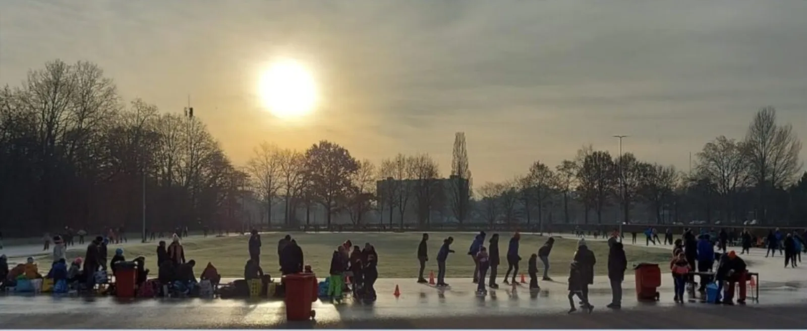 Schaatsers in silhouet op de Rubensbaan in de lage winterzon, met tassen en rode afvalbakken langs de baan en kale bomen erachter. – Rubensbaan (Amersfoortse IJsvereniging)