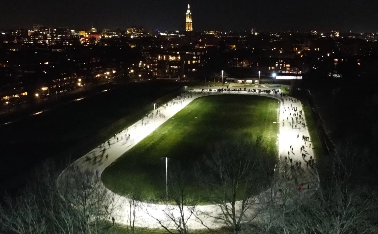 Een dronebeeld in de nacht van de verlichte Rubensbaan in Amersfoort vol schaatsers op de ovale ijsbaan rond het grasveld, met de lichten van de stad en de kerktoren erachter. – Rubensbaan (Amersfoortse IJsvereniging)