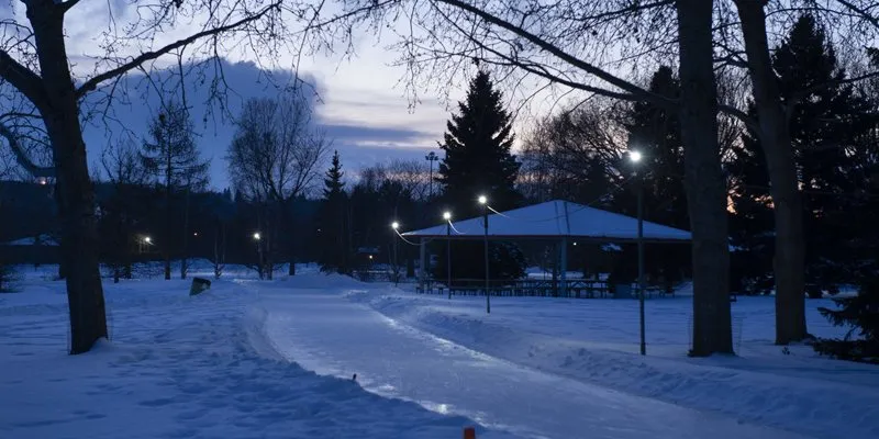 Rundle Park IceWay al atardecer, un sendero de patinaje iluminado entre árboles nevados, junto al pabellón del parque, sin personas a la vista (foto de la Ciudad de Edmonton) — Rundle Park IceWay and Shinny Ice Surface