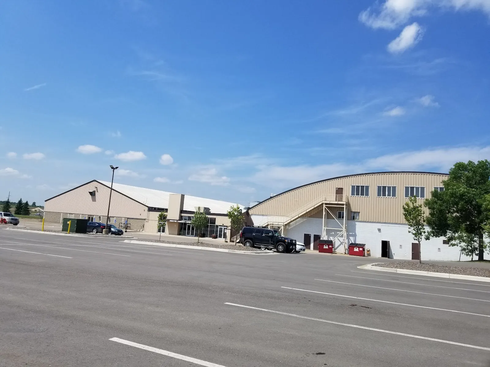 Front of the tan metal-sided building from the parking lot on a clear day, showing the main entrance with 'Runestone Community Center' signage, a black SUV and a few other cars parked nearby, and the taller arched roofline of the rink wing behind the entrance block. — Runestone Community Center