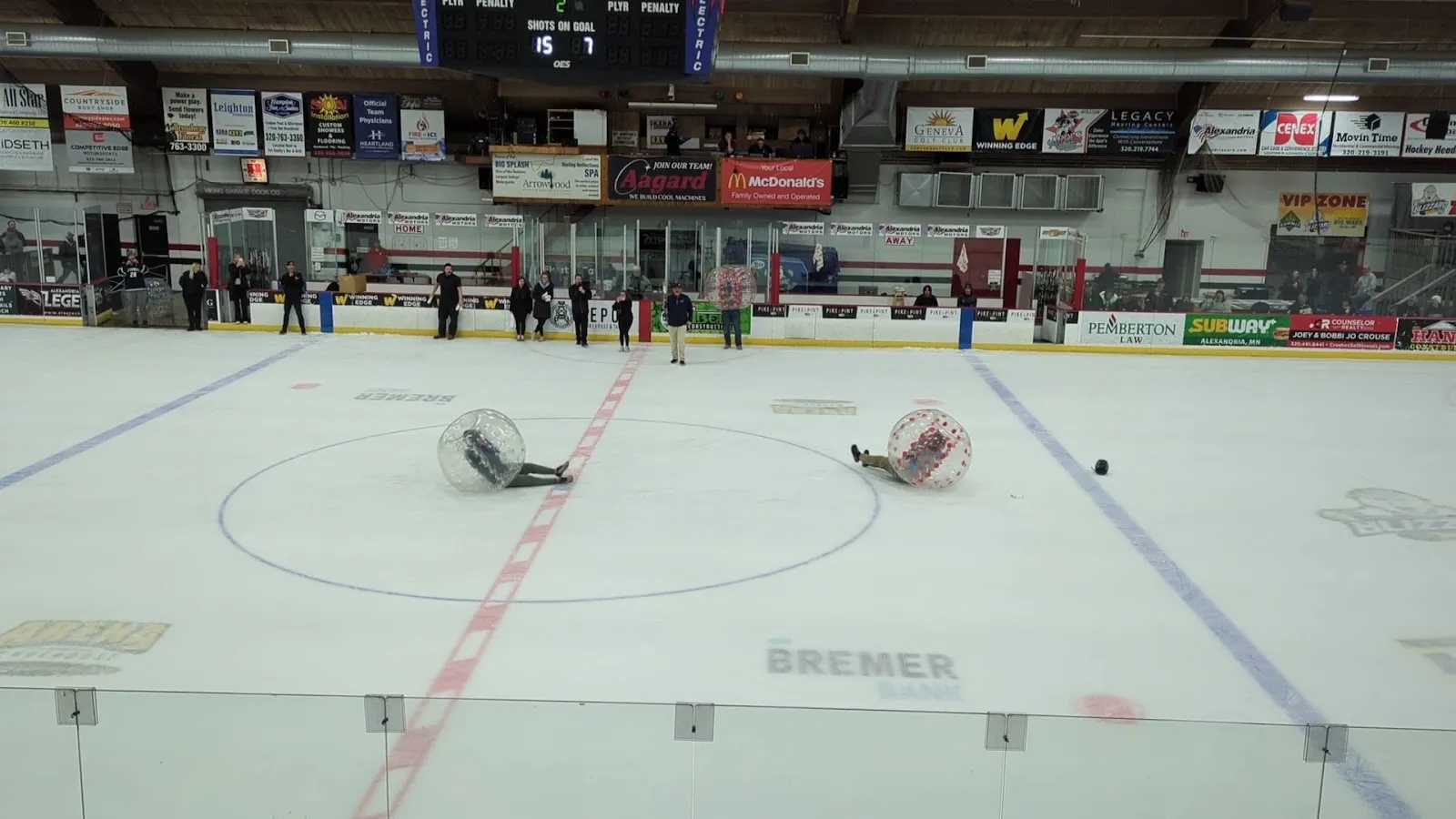 Players spread at centre ice for a faceoff with two inflatable mascots lying on the ice, seen through the glass under a timber roof with banners along the far wall. — Runestone Community Center