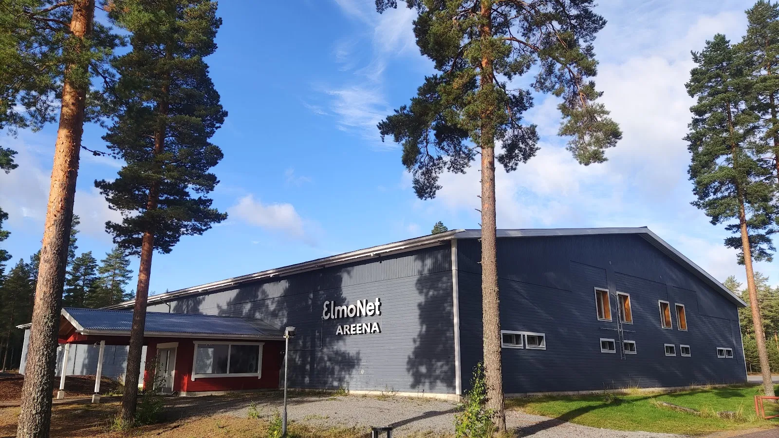 The dark blue-grey Ruovesi ice hall behind tall pines under a clear sky. – Ruoveden jäähalli