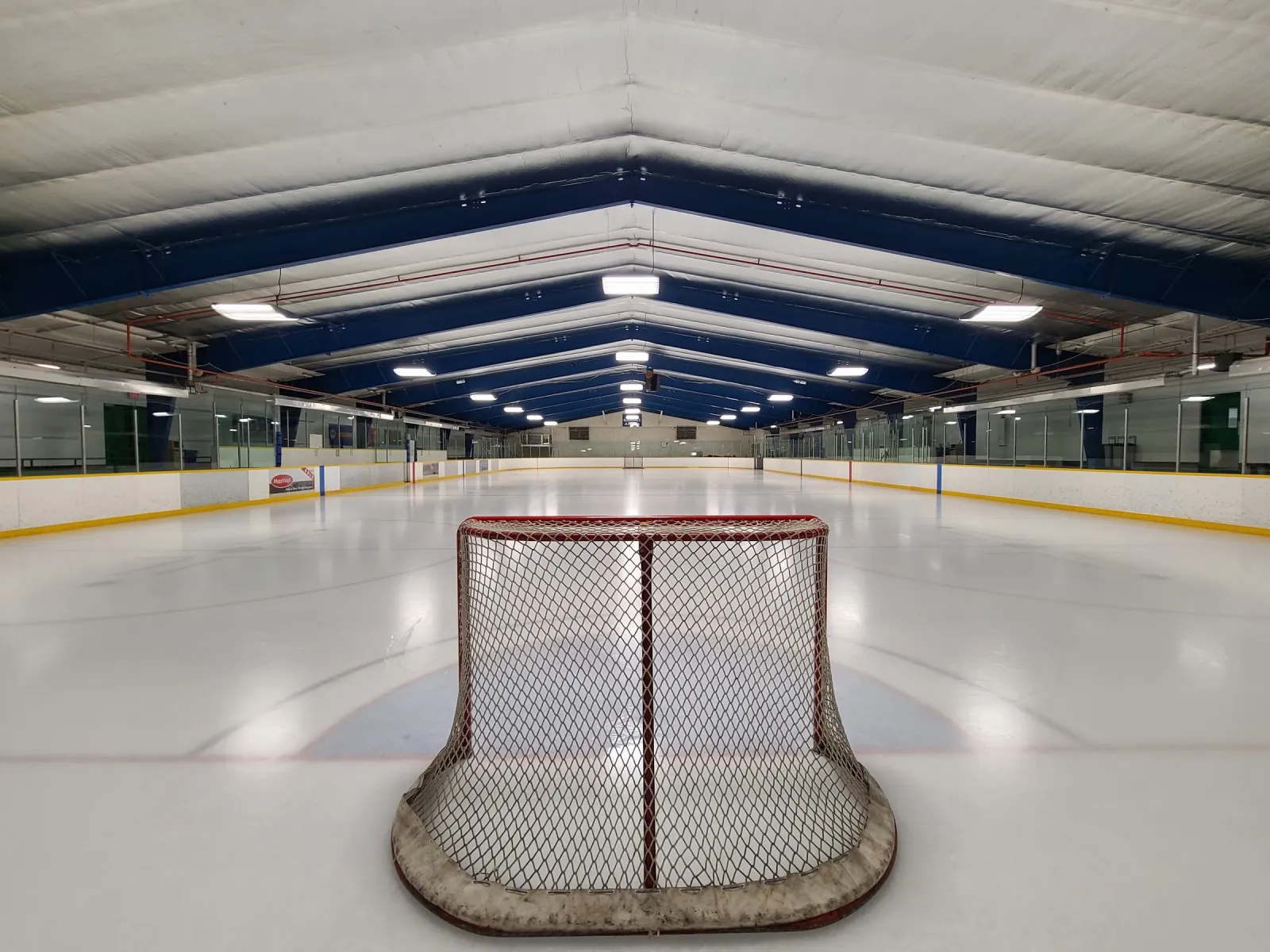 El hielo vacío visto desde detrás de una portería, a todo lo largo de la pista bajo un techo blanco con vigas azules. — Russ Barnes Arena