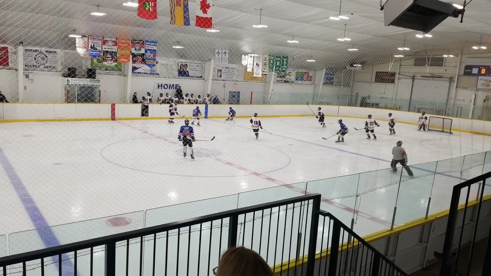 A wide view of a game from the rail, players spread across a white sheet with a banner of flags and a long upper wall of pale panels behind. — Frank Kinnaird Russell Arena