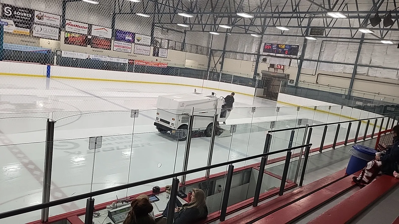An ice resurfacer on the sheet, seen from the red-and-white bleachers, with sponsor boards and flags on the far wall under blue roof trusses. — Rutland Arena
