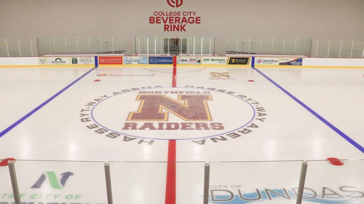 Centre ice of the new rink: the Northfield Raiders N inside the arena's name ring, red and blue lines, and sponsor boards below the far-end glass. — Ryt-Way Arena