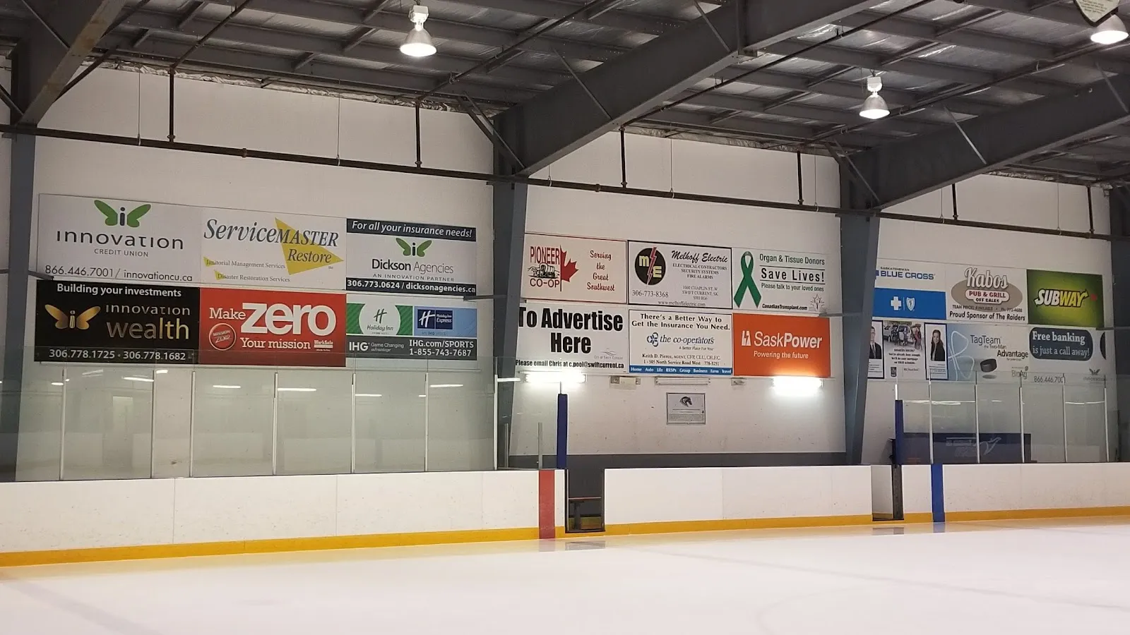 Una pista vacía en S3 Arenas: hielo limpio frente a las vallas y el vidrio, letreros de patrocinadores locales en el muro del extremo y un techo de armaduras de acero — S3 Arenas