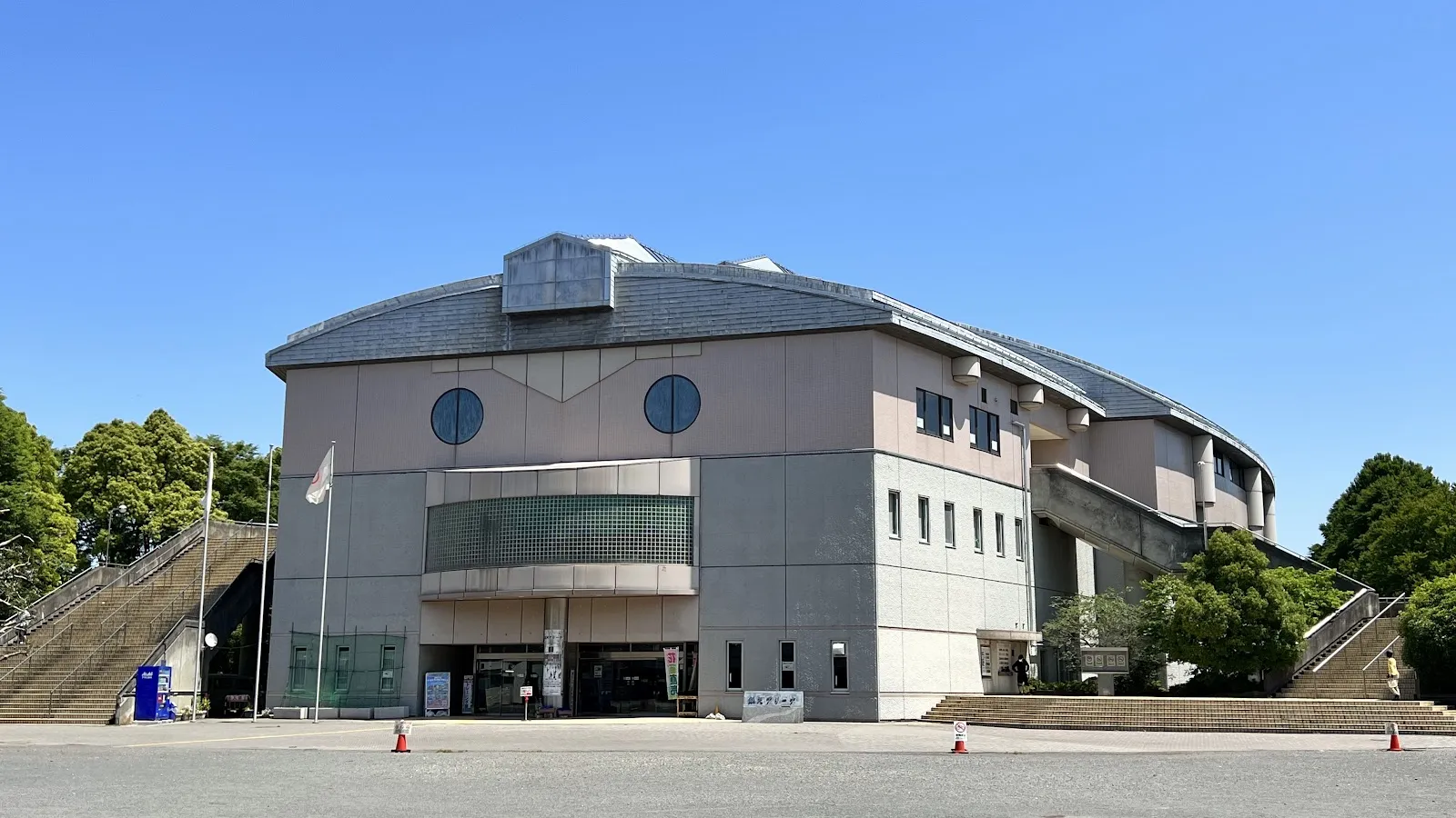 The grey and pink Sagamihara Ginga Arena building under a clear blue sky, with steps climbing on the left and the entrance below. — Sagamihara Ginga Arena