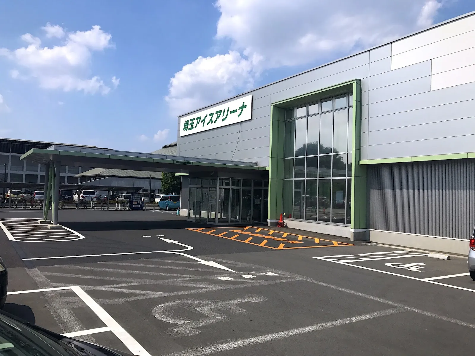 The front of Saitama Ice Arena in Ageo seen from the car park, with the green sign reading 埼玉アイスアリーナ above the glass entrance — Saitama Ice Arena