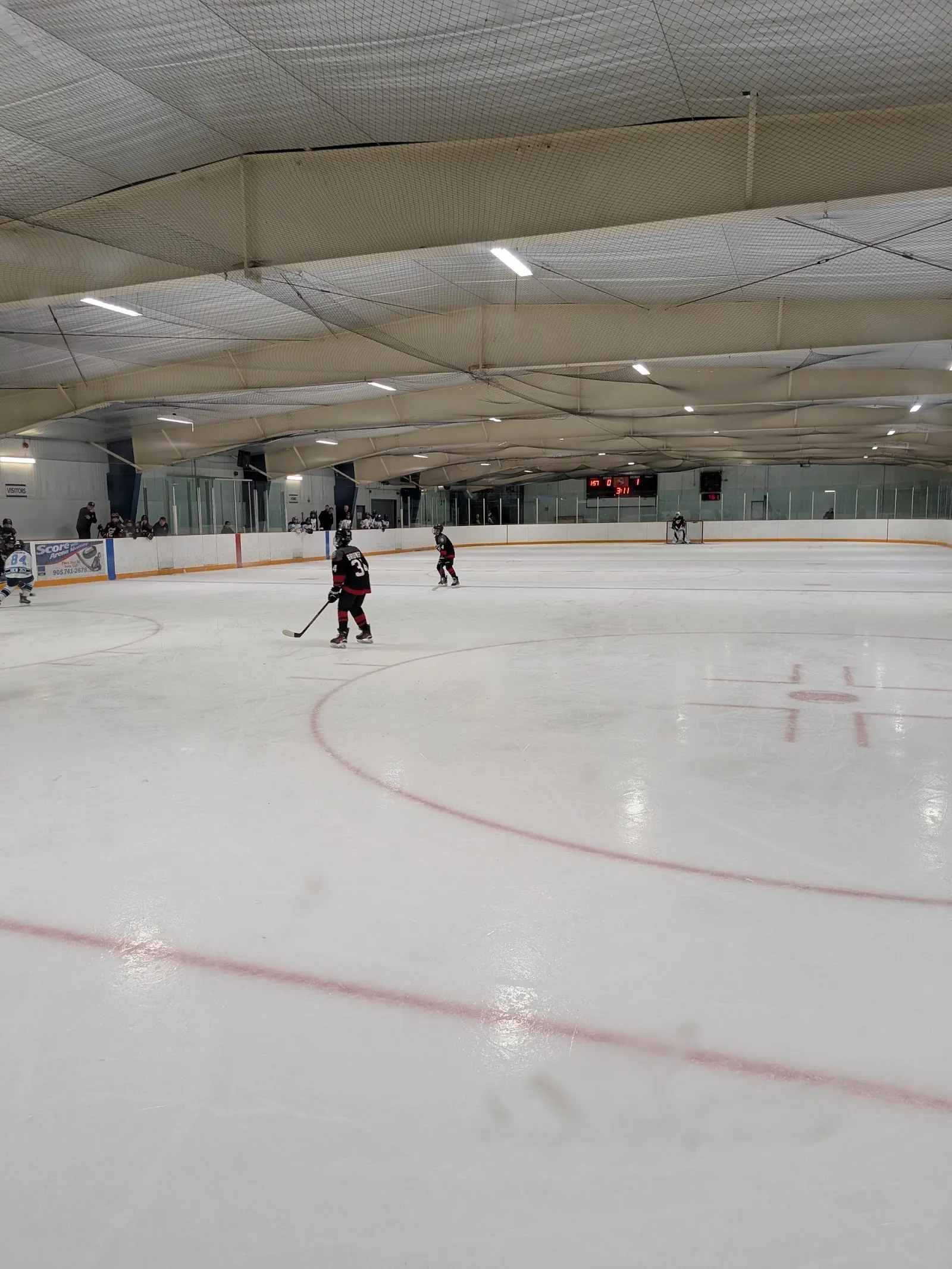 One skater on a wide sheet of white ice with red and blue lines and orange boards, a curved cream roof and rink glass beyond. — Saltfleet Arena