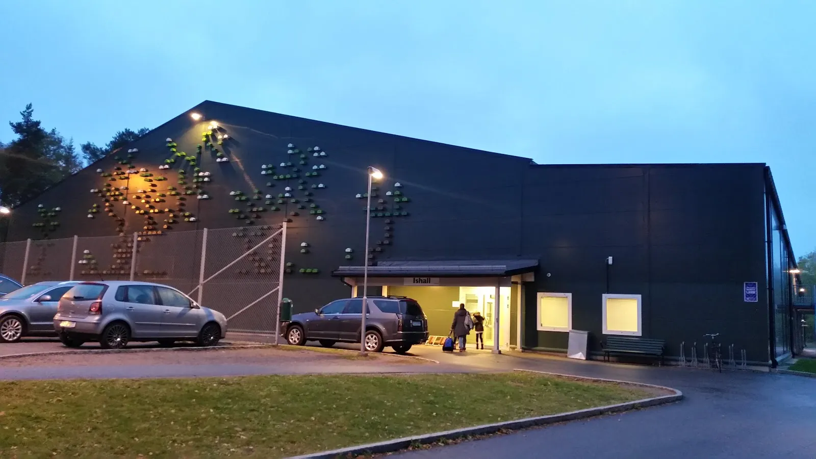 Den mörka Saltsjöbadens ishall i skymningen med ljusmönstret på gaveln och den upplysta entrén vid parkeringen – Saltsjöbadens ishall