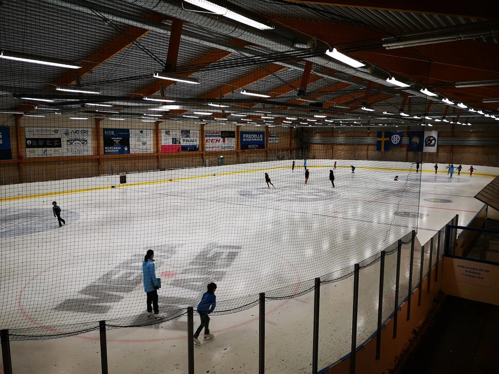 Skridskoåkare och spelare utspridda över isen i Saltsjöbadens ishall, sedda över nätet längs sargen under trätaket. – Saltsjöbadens ishall
