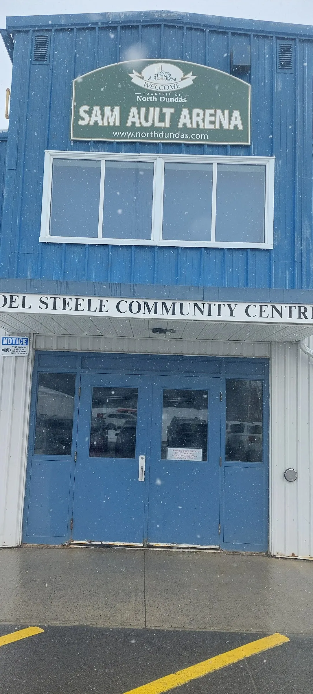 El frente del Sam Ault Arena y el Joel Steele Community Centre, un edificio metálico revestido de azul con el nombre de la arena en un letrero verde sobre un par de puertas azules. — Sam Ault Arena (Winchester)