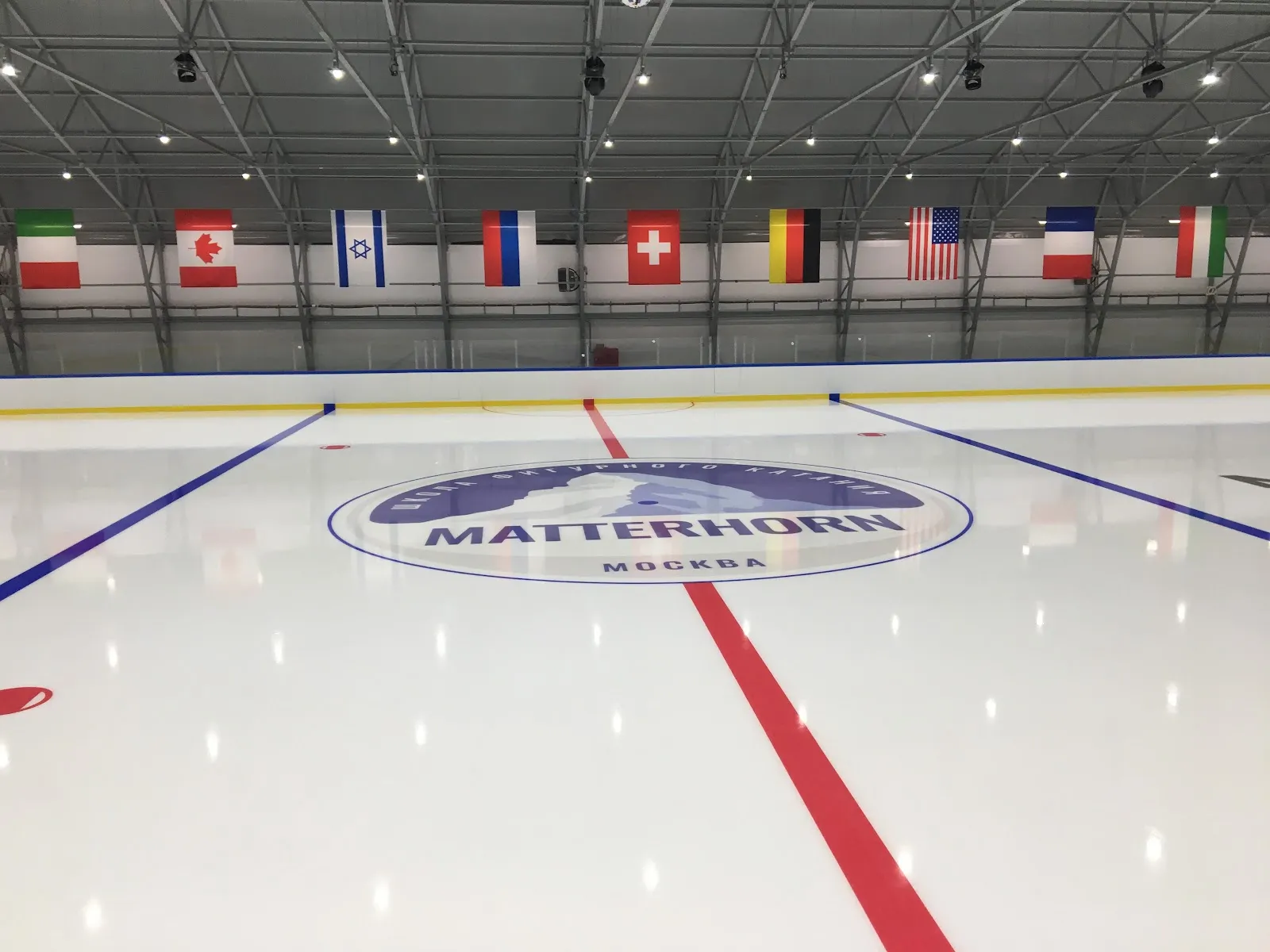 The ice sheet of the arena with the Matterhorn logo on the centre circle and flags along the wall — Samolet MIC Arena (Matterhorn)