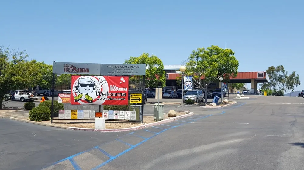 La entrada vehicular con el letrero rojo de bienvenida de San Diego Ice Arena y su mascota pingüino, con el estacionamiento y árboles detrás. — San Diego Ice Arena
