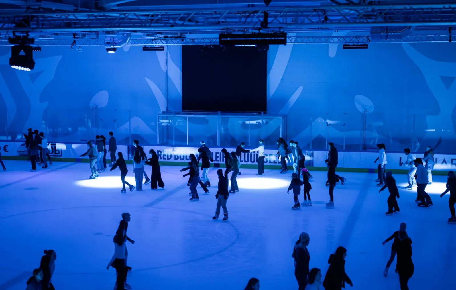 Eisläuferinnen und Eisläufer verteilen sich bei einer Eisdisco in blauem Licht auf einer der unterirdischen Eishallen, dahinter die Bande und eine Videowand. – SAP Garden Eispark