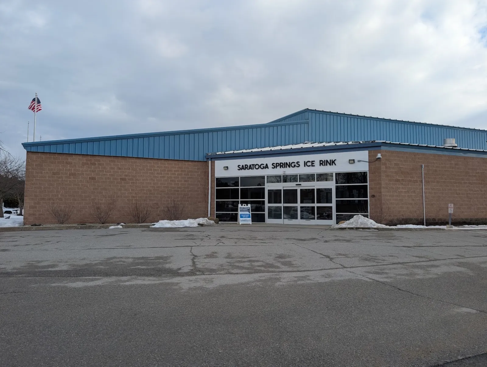 The low brick-and-blue-metal rink building behind its parking lot, with "Saratoga Springs Ice Rink" lettered over the glass entrance. — Saratoga Springs Ice Rinks