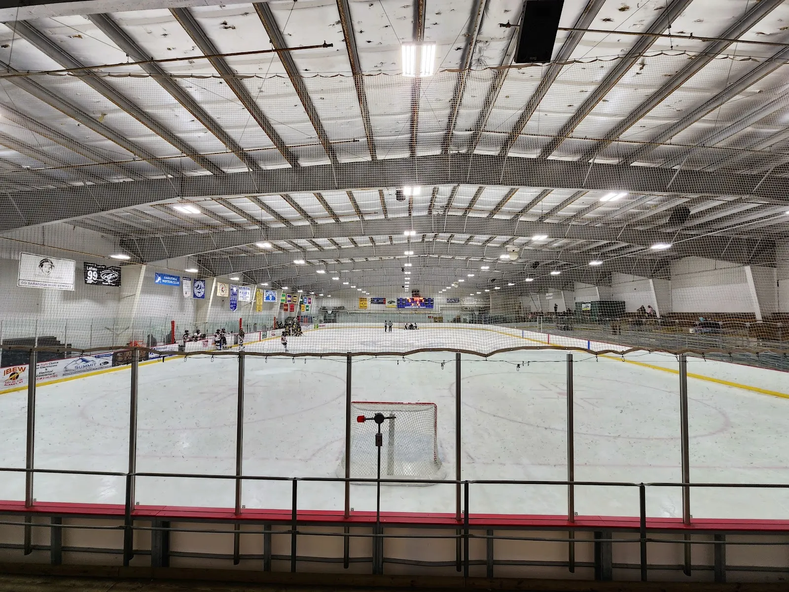 A full sheet seen from the end boards behind a net, under a long arched steel roof, with a few skaters far down the ice. — Saratoga Springs Ice Rinks