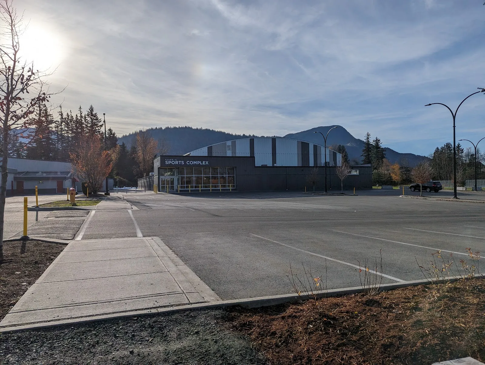 The Sardis Sports Complex entrance with its name above the glass doors, the curved arena roof behind and mountains beyond the parking lot. — Sardis Sports Complex