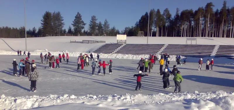 Children skating on the ice at the Särkkiranta sports centre skating field, with the grandstand along the far side. — Sarkkirannan urheilukeskuksen luistelukenttä (Kempele)