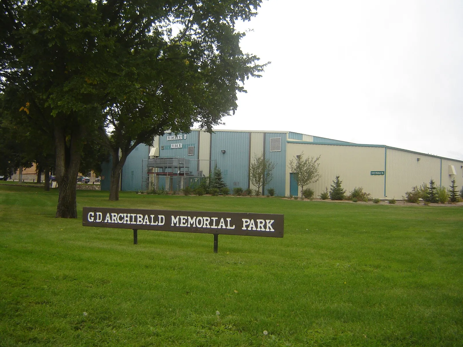 The G.D. Archibald Memorial Park sign on the lawn, with the long blue-and-white Archibald Arena building behind the trees. — Archibald Arena