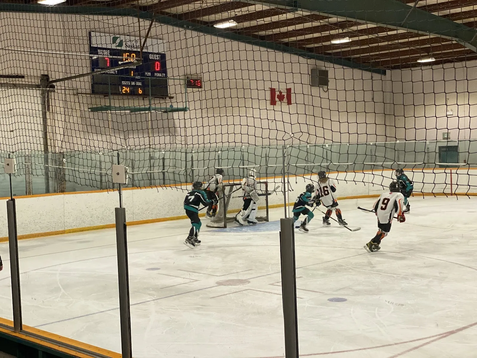 A game seen across a netted corner, players spread across the sheet under a timber and steel roof with a Canadian flag and scoreboard on the far wall. — Archibald Arena