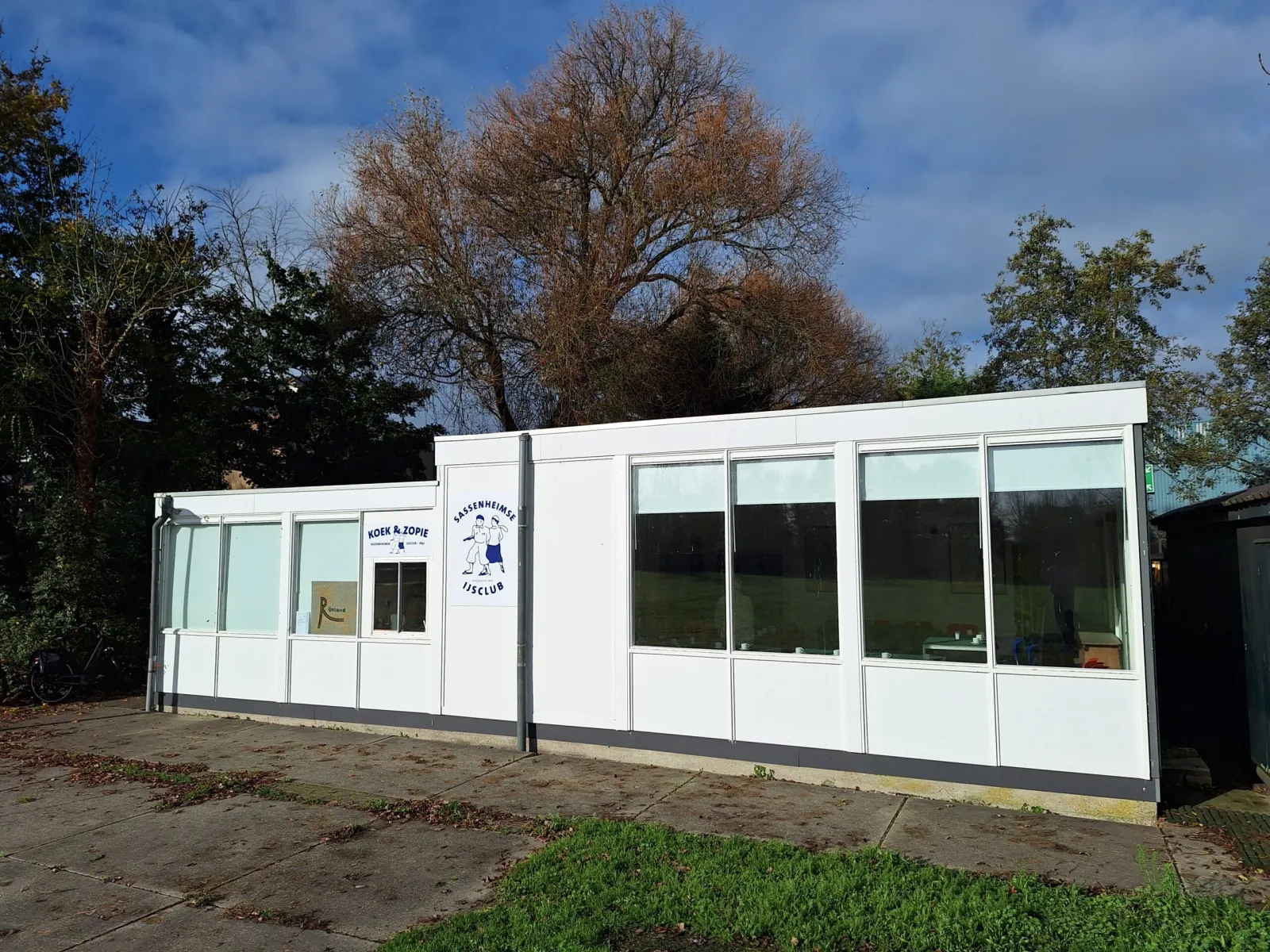 The white clubhouse of the Sassenheimse IJsclub with a koek-en-zopie hatch and the club emblem, seen from the paved area in front under a blue sky with an autumn tree behind. — Landijsbaan Sassenheim (Sassenheimse IJsclub)