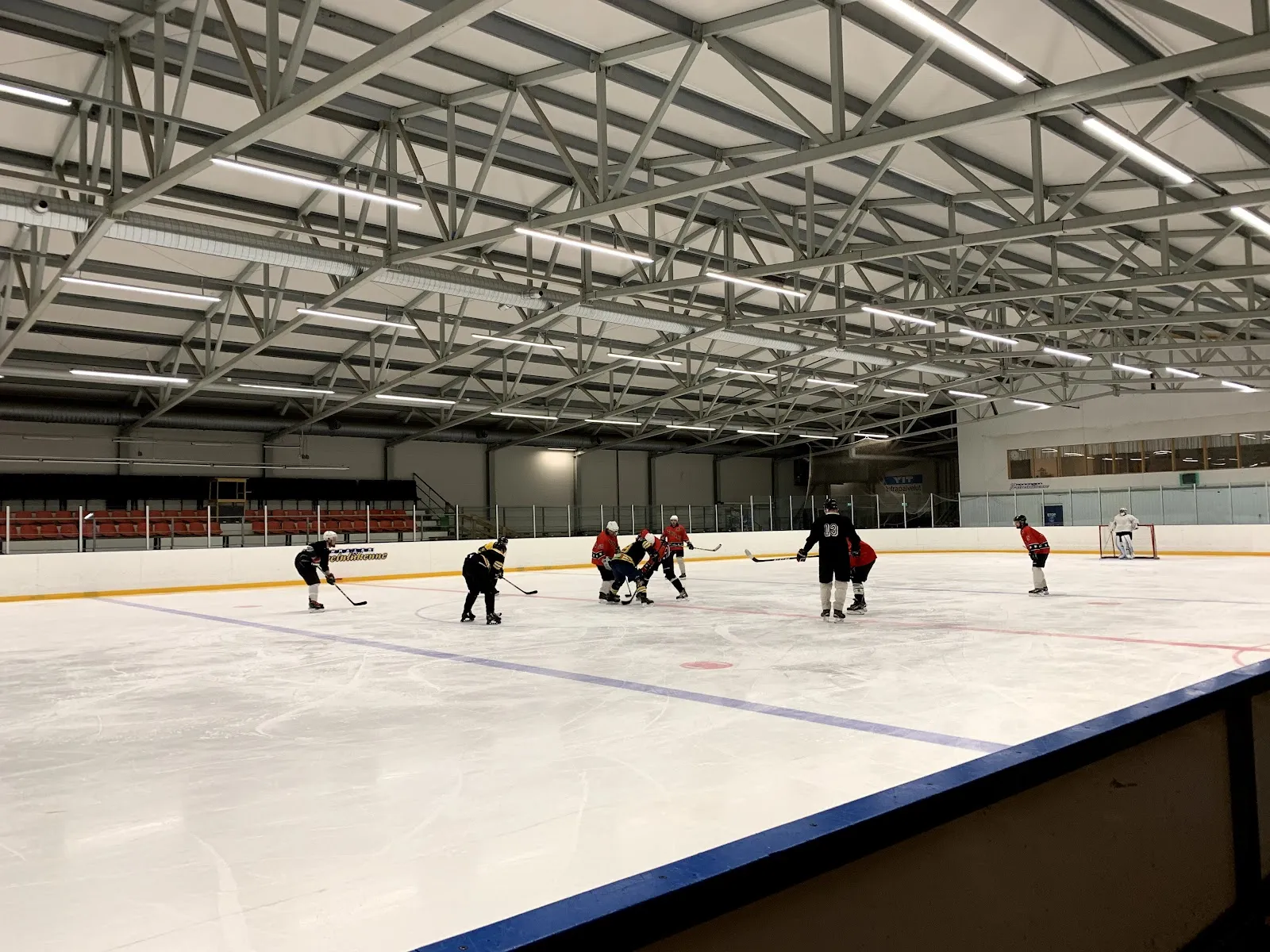A wide view along the Sastamala ice hall rink with a few players far down the ice and the blue boards in the foreground. — Sastamalan jäähalli