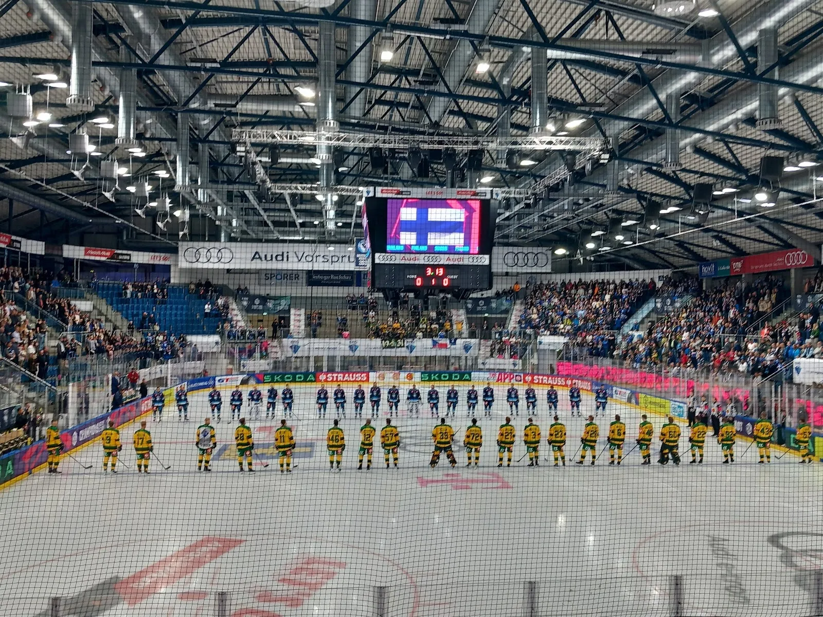 The ice sheet of the SATURN-Arena in Ingolstadt before a match, with the two teams lined up along the blue line and the stands, scoreboard and board advertising behind. — SATURN-Arena und Zweite Eishalle