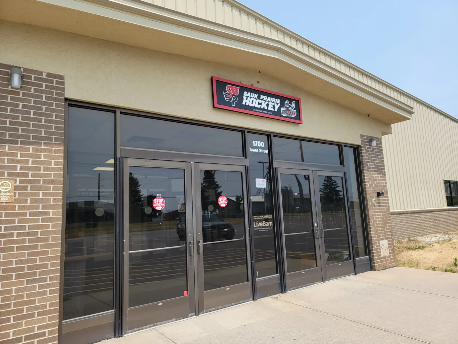The rink's glass double-door entrance in a brick-and-tan-metal building, with a Sauk Prairie Hockey sign above and the street number 1700 on the glass. — Sauk Prairie Ice Arena
