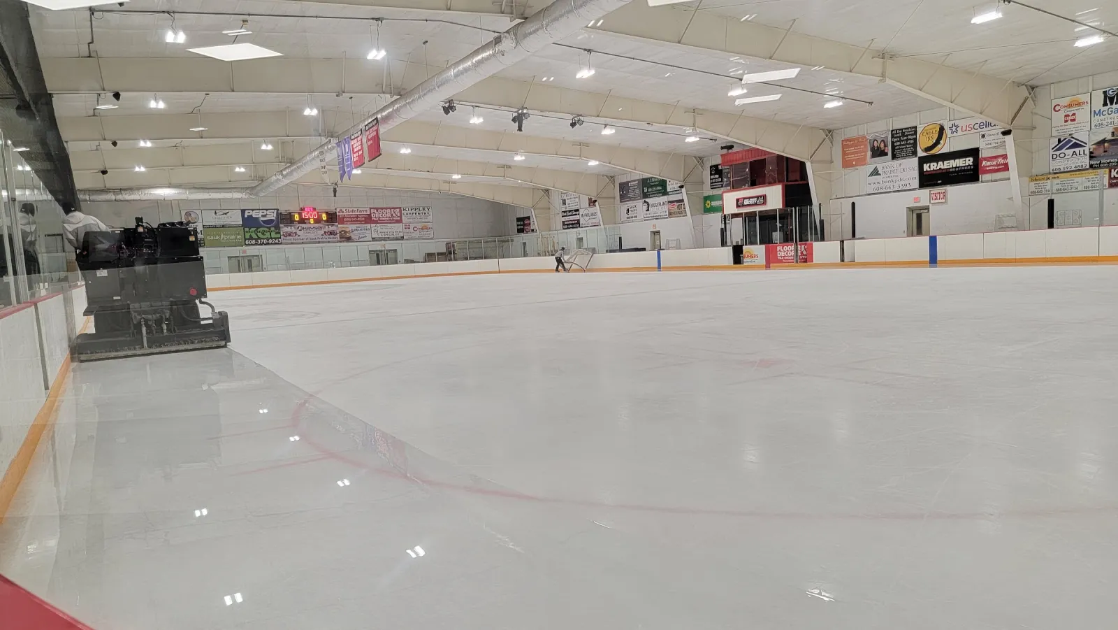 The empty rink at Sauk Prairie Ice Arena, with the ice resurfacer parked by the boards and sponsor signs along the far wall. — Sauk Prairie Ice Arena
