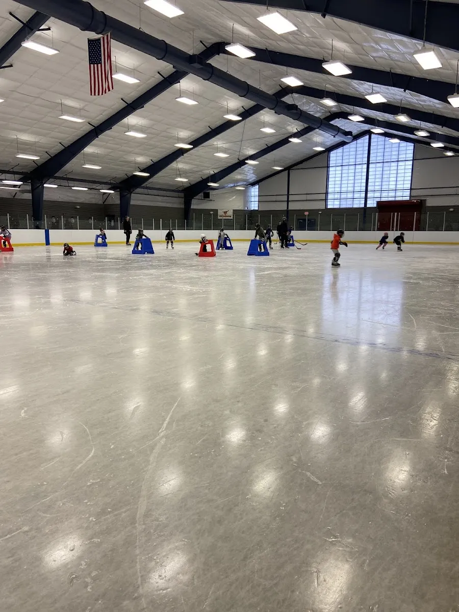 The wide, empty ice surface of the Scanlon indoor rink under the roof lights, with boards and a few skaters in the distance. — Scanlon Ice Rink