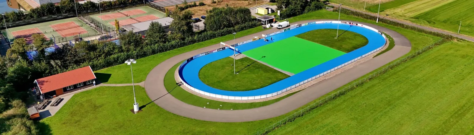 Aerial view of the Rijssen skating site: the blue-edged 200 m inline oval with its green infield, the flat asphalt track around it, the red-roofed clubhouse and tennis courts beside it. — Natuurijsbaan Rijssen (Schaats- en Skeelervereniging Rijssen)