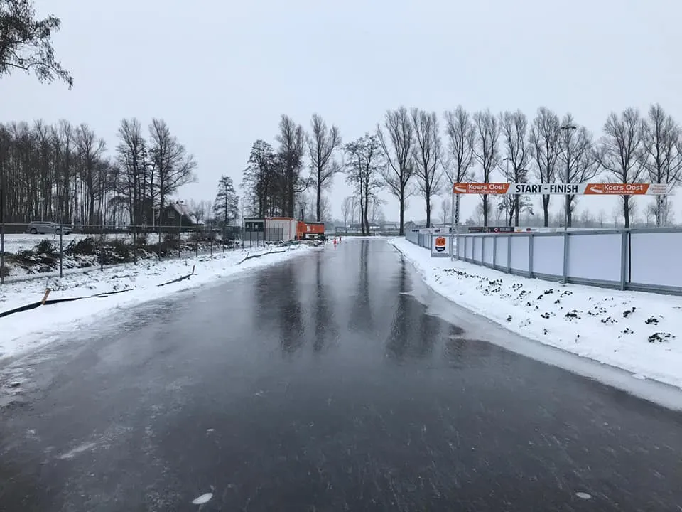 The asphalt track at Rijssen covered in smooth natural ice in winter, with snow along the edges, bare poplars and the start and finish gantry in the distance. — Natuurijsbaan Rijssen (Schaats- en Skeelervereniging Rijssen)