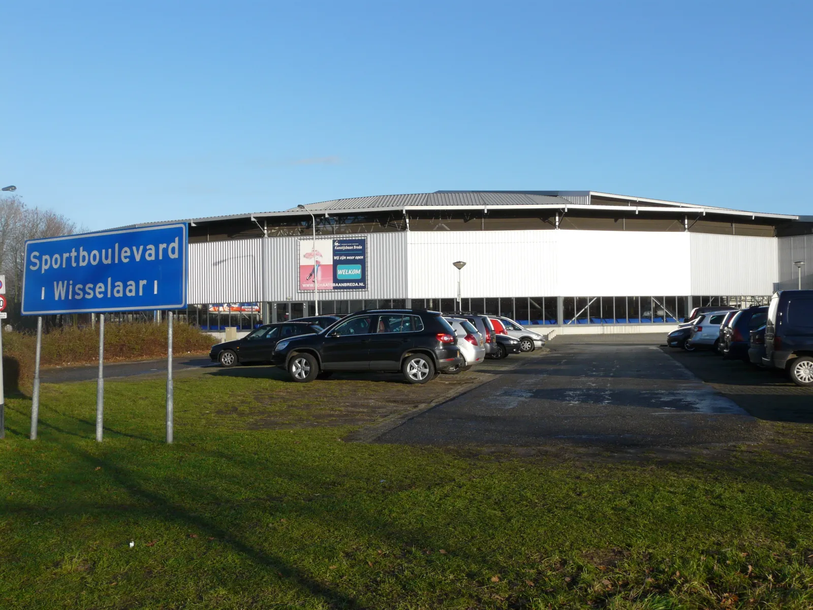 De Kunstijsbaan Breda aan Sportboulevard Wisselaar in Breda gezien vanaf het parkeerterrein, een lang wit en grijs gebouw onder een plat dak met een blauw verkeersbord ervoor – Schaatsbaan Breda