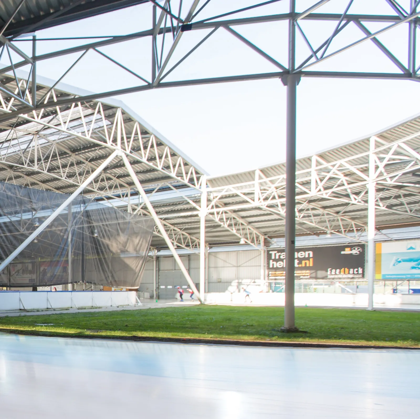 De overdekte 400 m-baan van Schaatsbaan Breda, bleek ijs dat langs een grasmidden buigt onder de witte stalen dakspanten, met schaatsers ver weg op de achterbocht – Schaatsbaan Breda