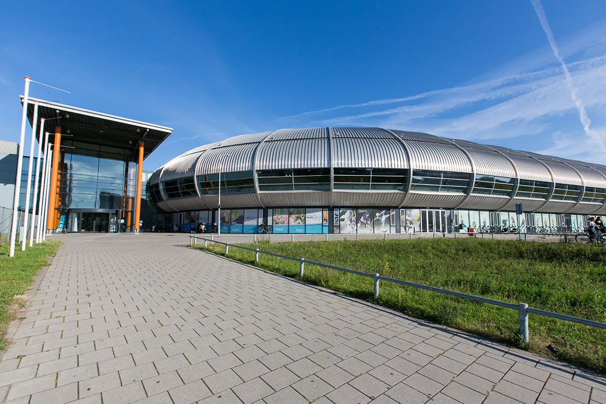 The Sportboulevard Dordrecht building from the paved forecourt, a glass entrance with an orange frame beside the ribbed silver curved wall of the ice hall under a blue sky — Schaatsbaan Dordrecht