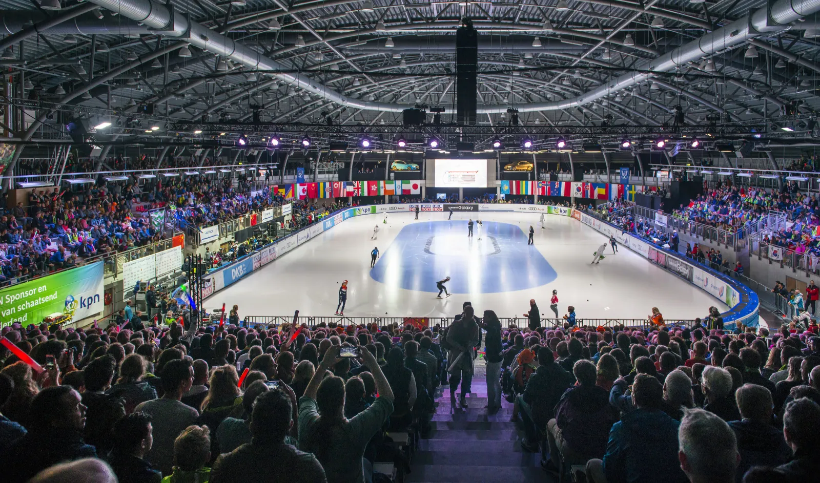 The ice hall of Sportboulevard Dordrecht during a short-track World Cup, the white rink seen from the stands with a crowd of spectators with their backs to the camera and national flags along the far side — Schaatsbaan Dordrecht