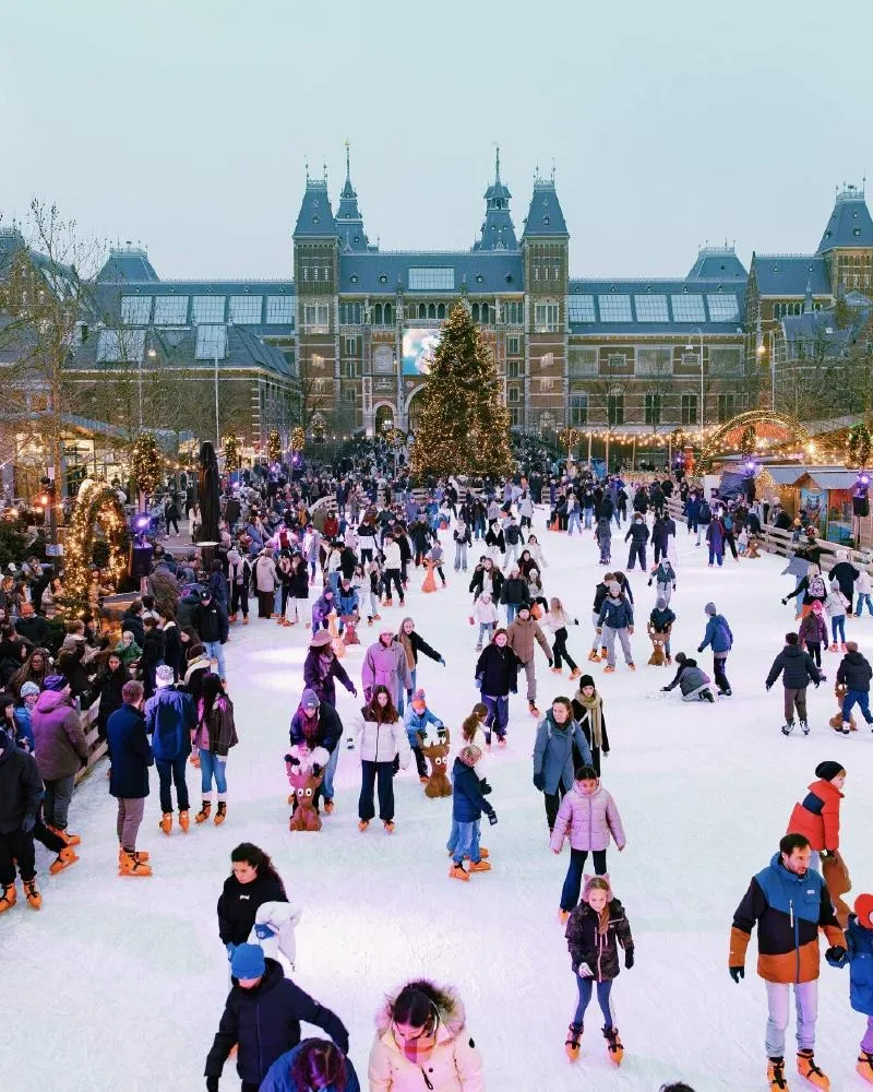 De schaatsbaan op het Museumplein voor het Rijksmuseum, een menigte schaatsers op het ijs met een kerstboom onder de museumgevel. – Schaatsbaan Museumplein