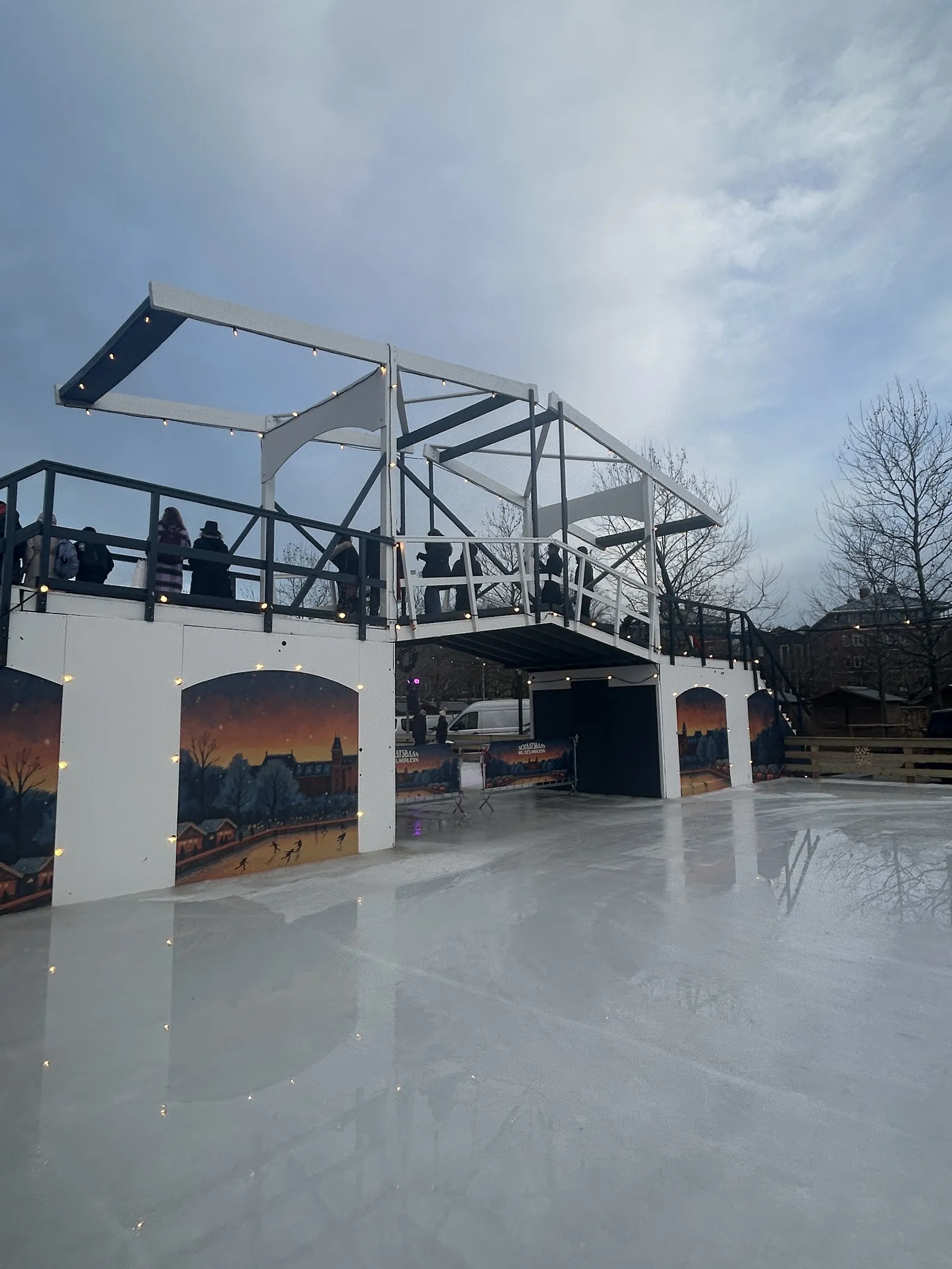 Het lege ijs van de schaatsbaan op het Museumplein onder een grijze lucht, met de witte uitkijkbrug en haar beschilderde bogen met winterse taferelen aan de overkant – Schaatsbaan Museumplein