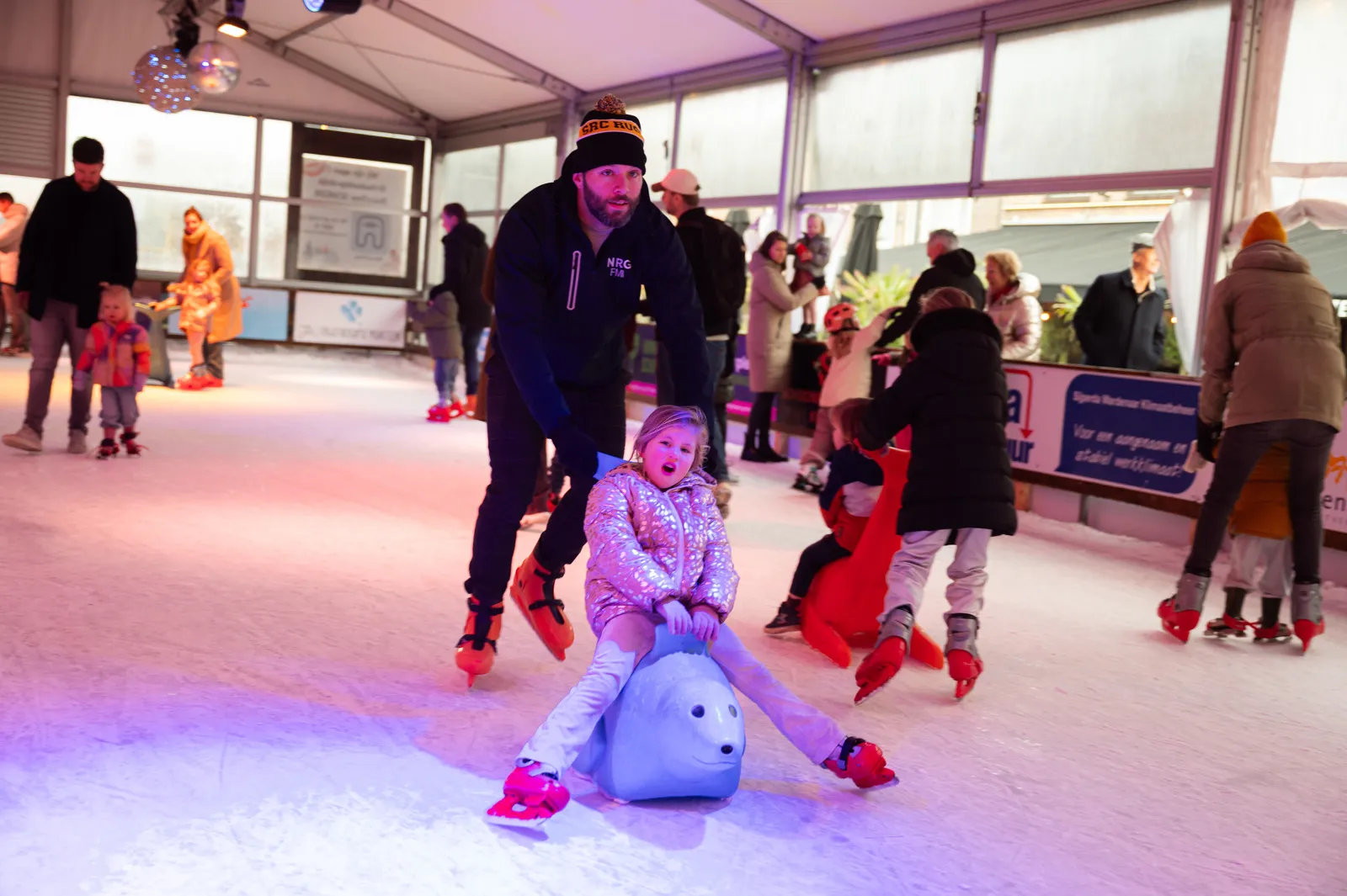 Kinderen op zeehondvormige schaatshulpen met een begeleidende volwassene op de overdekte ijsbaan van Schagen on Ice; schaatsers in actie, niemand poseert. – Schagen on Ice